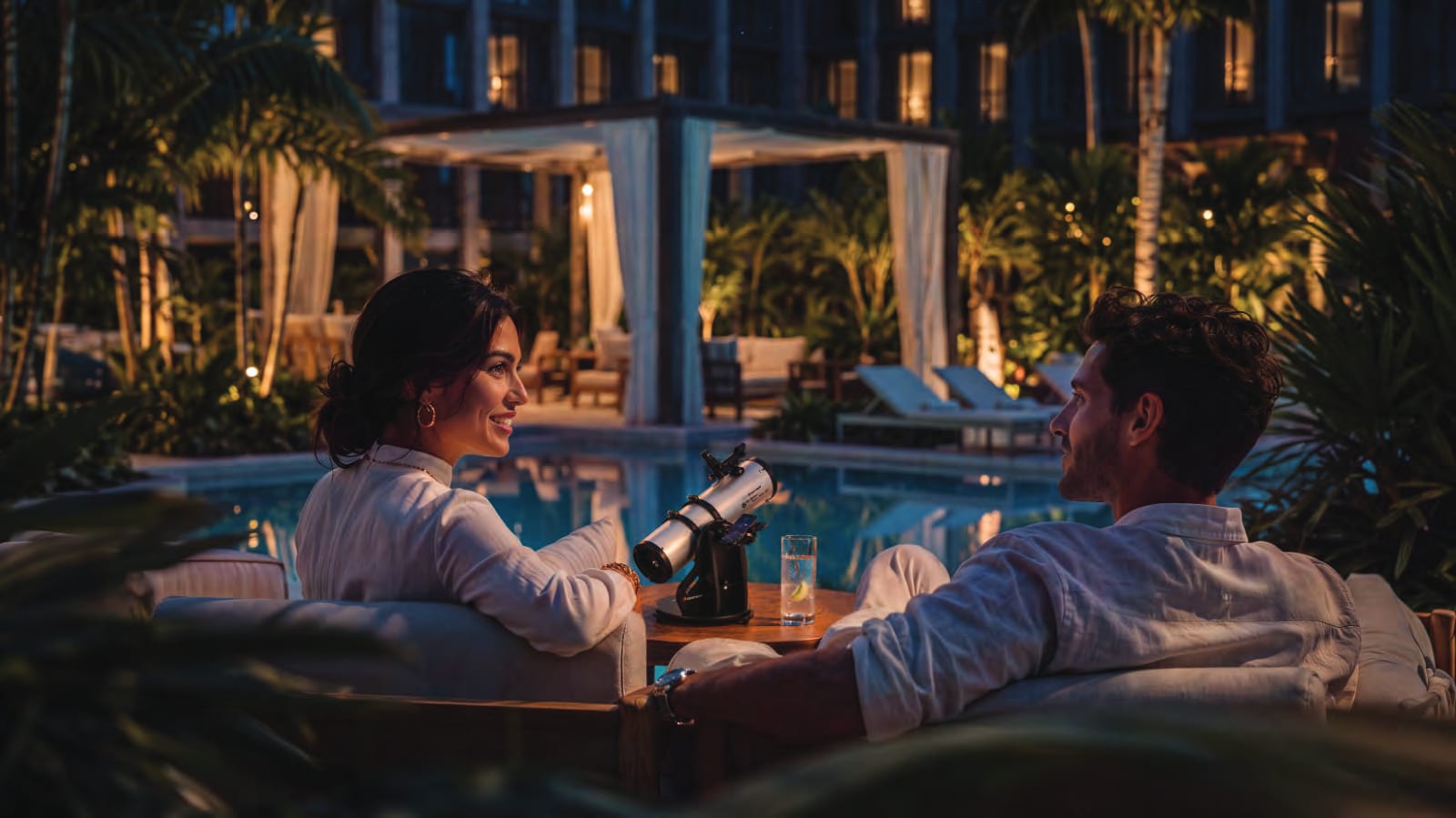 Two residents on a poolside sofa at night with a telescope on the table, beside a lit cabana on the podium