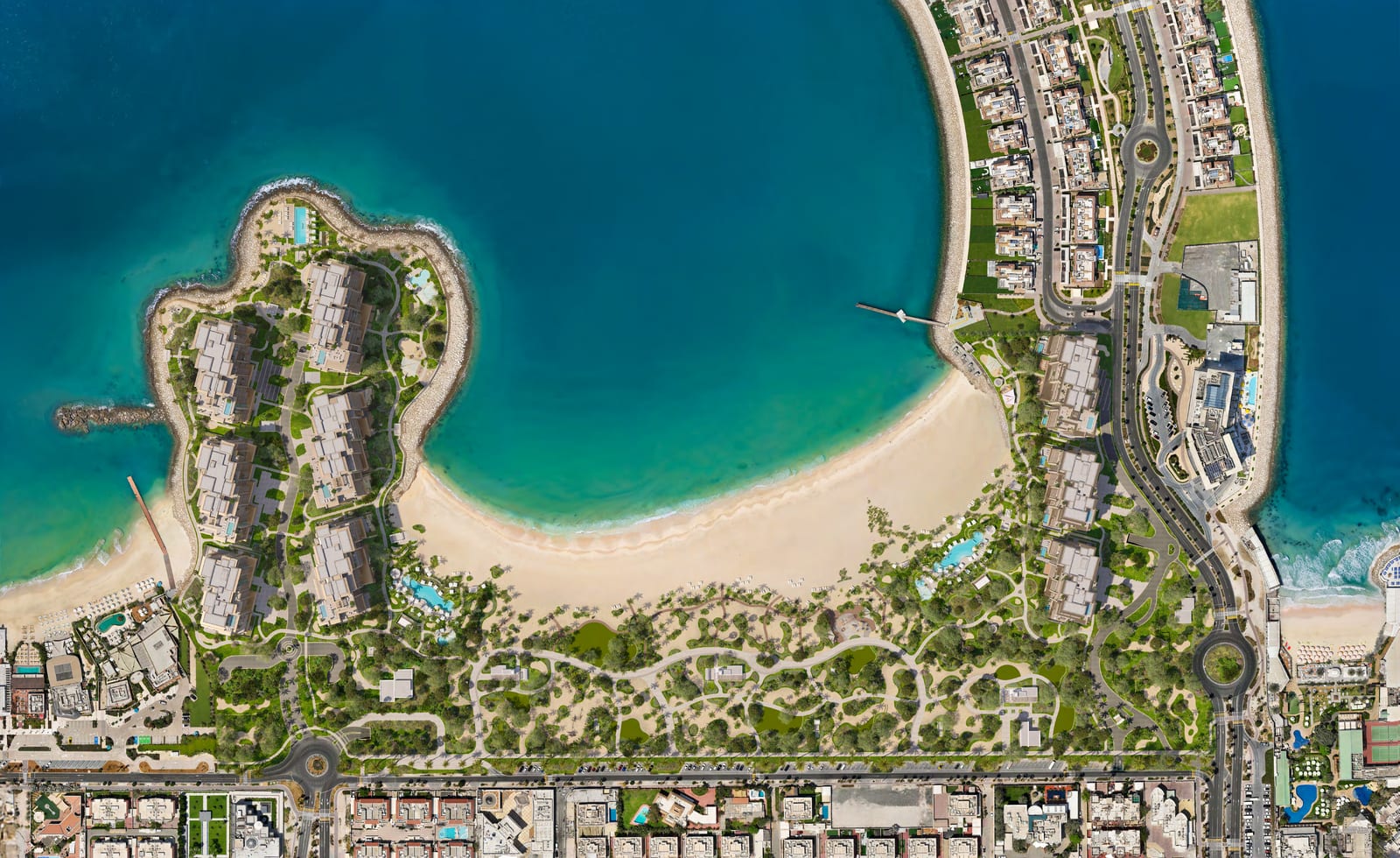 Aerial view of a crescent beach and turquoise bay, with six apartment buildings on a landscaped headland to the left and three more along the right-hand shore